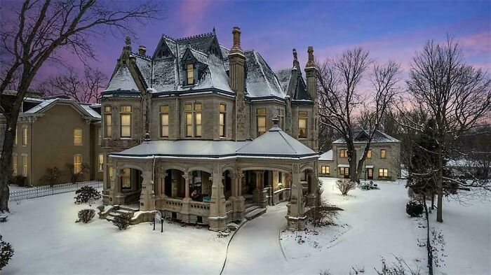 Charming old home covered in snow at dusk with intricate architecture and bare winter trees surrounding the property.