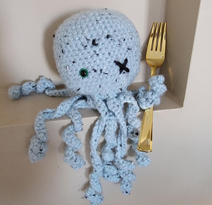Crochet animal with personality holding a gold fork with curly tentacles and one green eye displayed on a shelf.