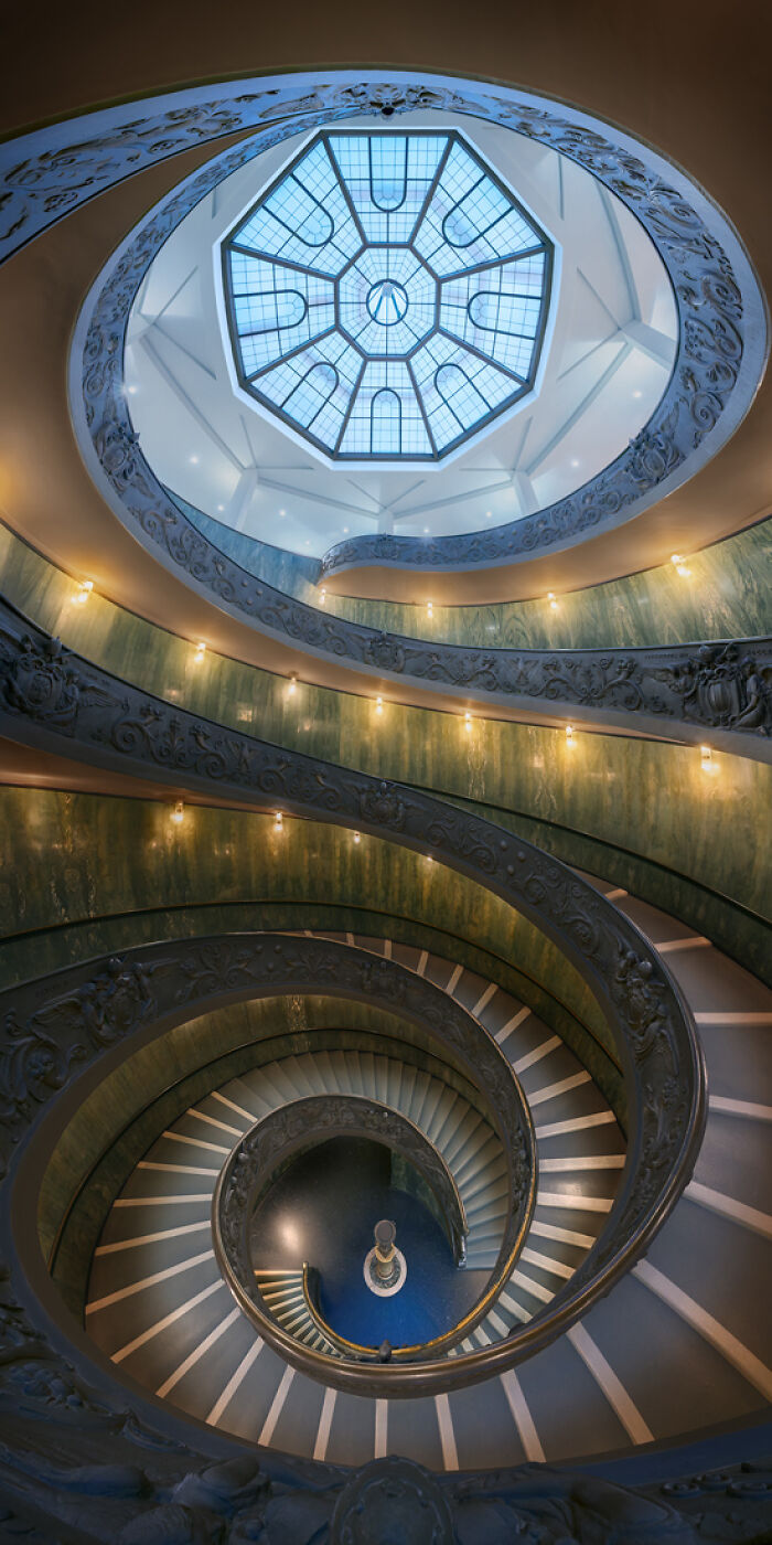 Spiral staircase with ornate railings and blue-tinted glass ceiling captured in an award-winning panoramic masterpiece