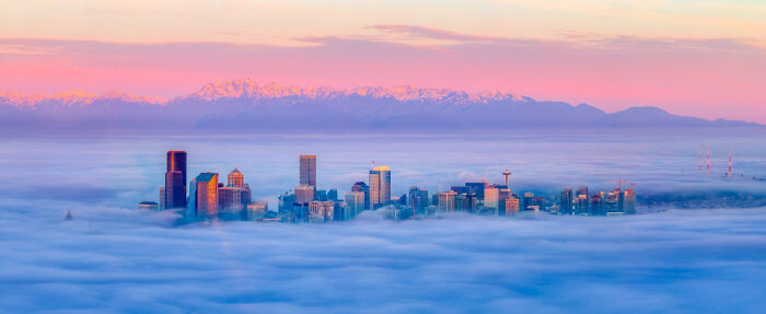 Panoramic cityscape of skyscrapers emerging through thick fog with mountains glowing under a pink and purple sunrise sky.