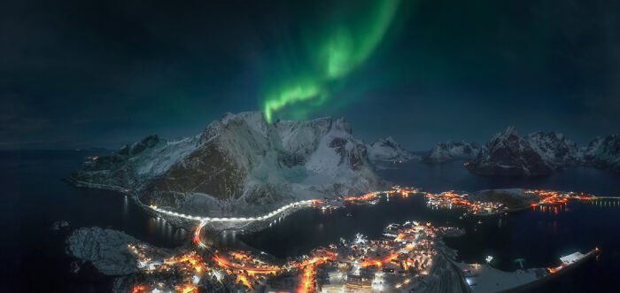 Northern lights over a snowy mountainous coastal village at night, showcasing panoramic masterpieces from Epson Pano Awards.