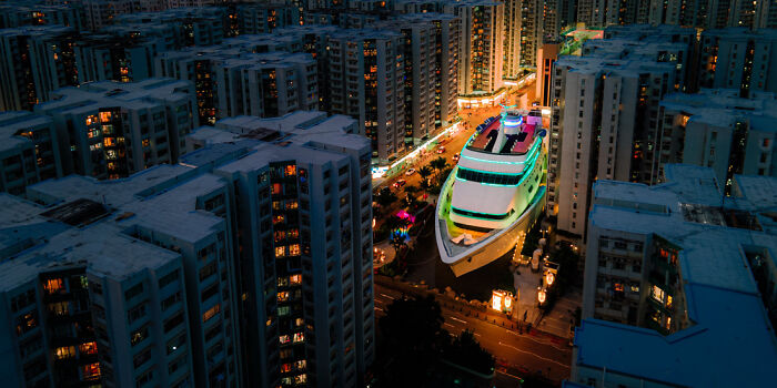 Panoramic masterpiece of a large illuminated yacht navigating through a narrow city street between tall buildings at night.