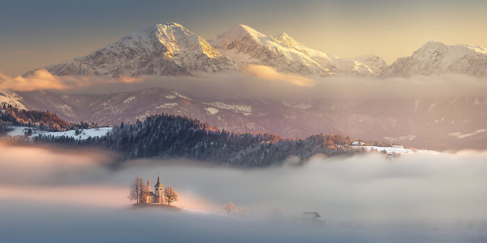 Panoramic masterpiece of snow-covered mountains and a church surrounded by mist during a golden sunrise.