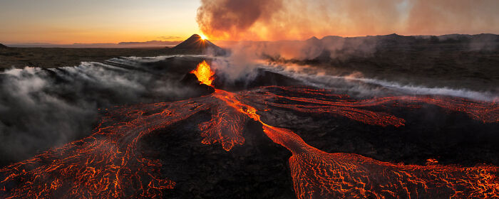 Panoramic masterpiece showing an active volcano erupting with flowing lava under a dramatic sunset sky.