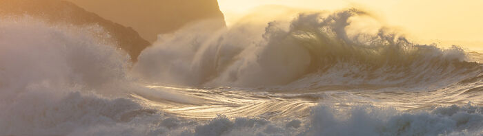 Panoramic masterpiece of powerful ocean waves crashing near rocky cliffs during a golden sunset, showcasing dynamic natural beauty.