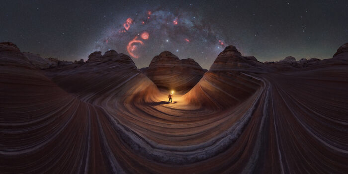 Panoramic landscape of wave-like rock formations under a starry sky with a person holding a light at the center.