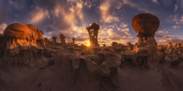 Panoramic masterpiece of a rocky desert landscape at sunset with dramatic clouds and sunburst lighting effects.