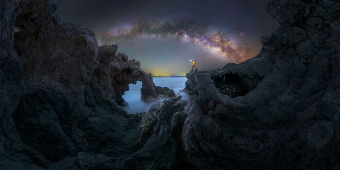Panoramic night landscape with a person capturing the Milky Way over rugged rocky formations and misty water.