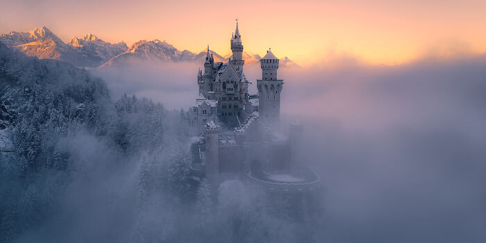 Panoramic view of a misty castle at sunrise with snow-covered trees and mountains in the background.