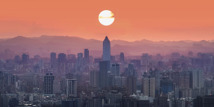 Panoramic cityscape at sunset with silhouetted skyline and layered mountains under a glowing sun, showcasing stunning panoramic masterpiece.