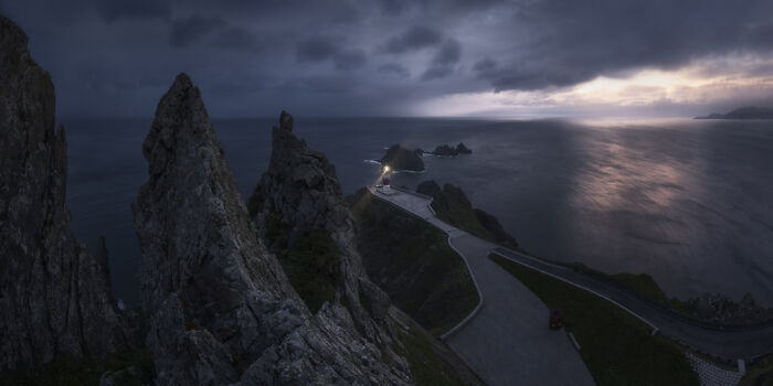 Dark panoramic landscape featuring jagged rocks and a winding road leading to a distant lighthouse at dusk, Epson Pano Awards.