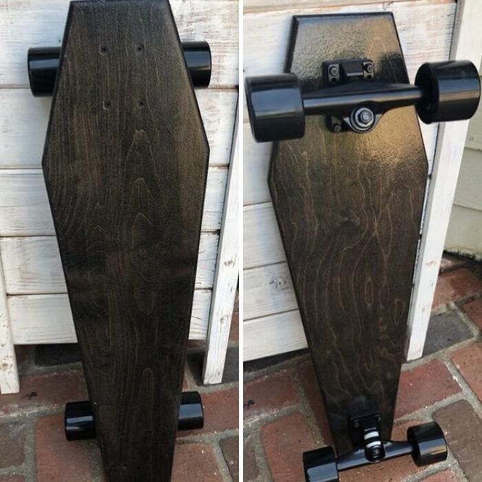 Black wooden coffin-shaped skateboard with black wheels leaning against a white wooden wall, perfect for horror and Halloween fans.