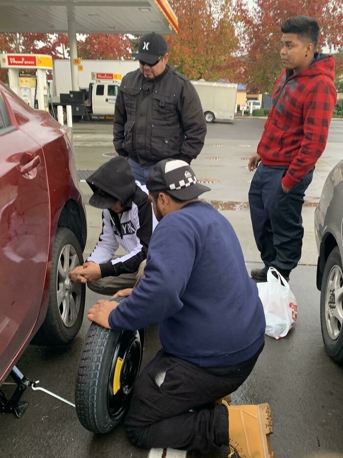 Strangers helping change a flat tire at a gas station, showing faith in humanity restored through kind actions.