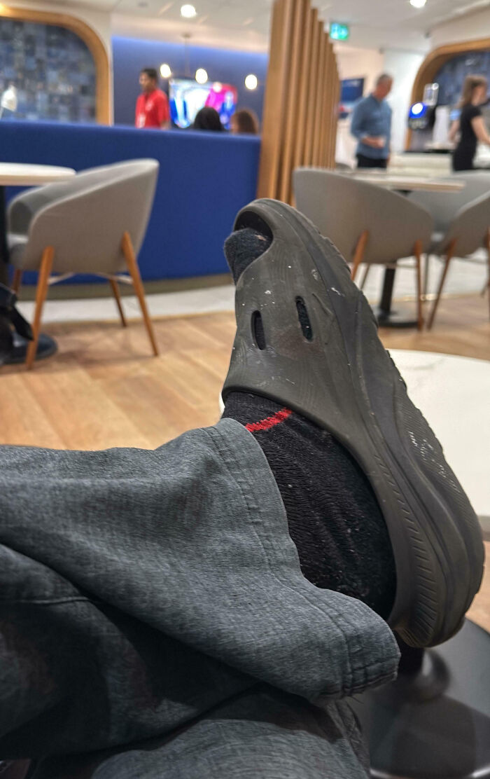 Close up of a worn black clog shoe and sock with casual pants in a busy indoor seating area, showing a messy moment.