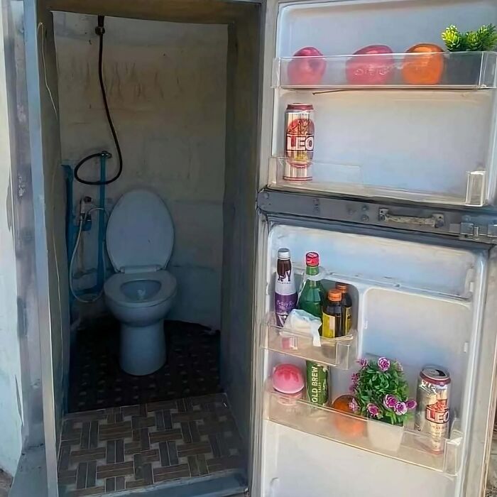 Open refrigerator door revealing food and drinks with a toilet visible through a small adjacent room in a strange setup.