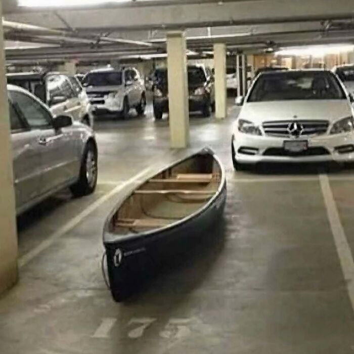 Canoe parked in a multi-level parking garage among cars, an unusual image that might need a priest or an exorcist.