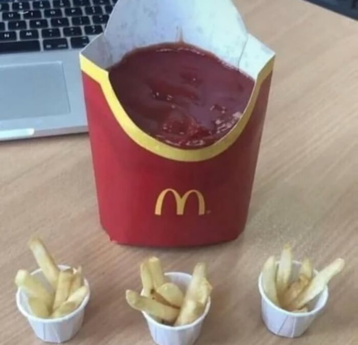Large McDonald's fries container filled with ketchup and three small cups, each with a few fries, on a wooden table.