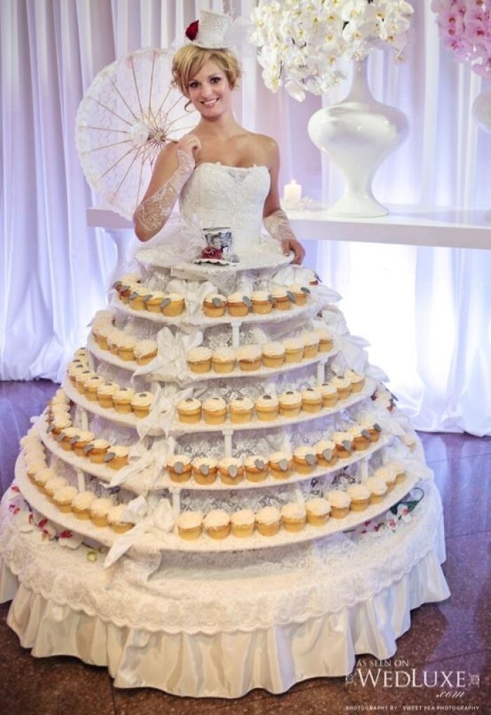 Woman wearing a wedding dress designed as a cupcake stand with multiple tiers holding cupcakes at a wedding event.
