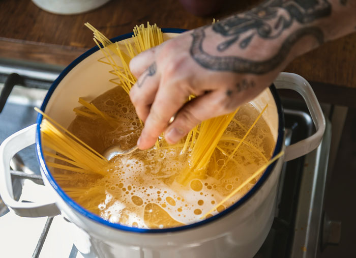 Hand with tattoos stirring uncooked spaghetti in boiling water, showcasing hilarious instances of people unaware of common knowledge.