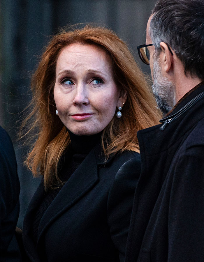 J.K. Rowling in a black coat with pearl earrings, engaging in conversation with a man wearing glasses and a dark jacket.