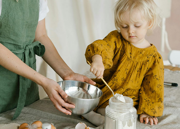 Young child in yellow dress scooping flour from jar while adult holds metal mixing bowl, demonstrating life hacks use.