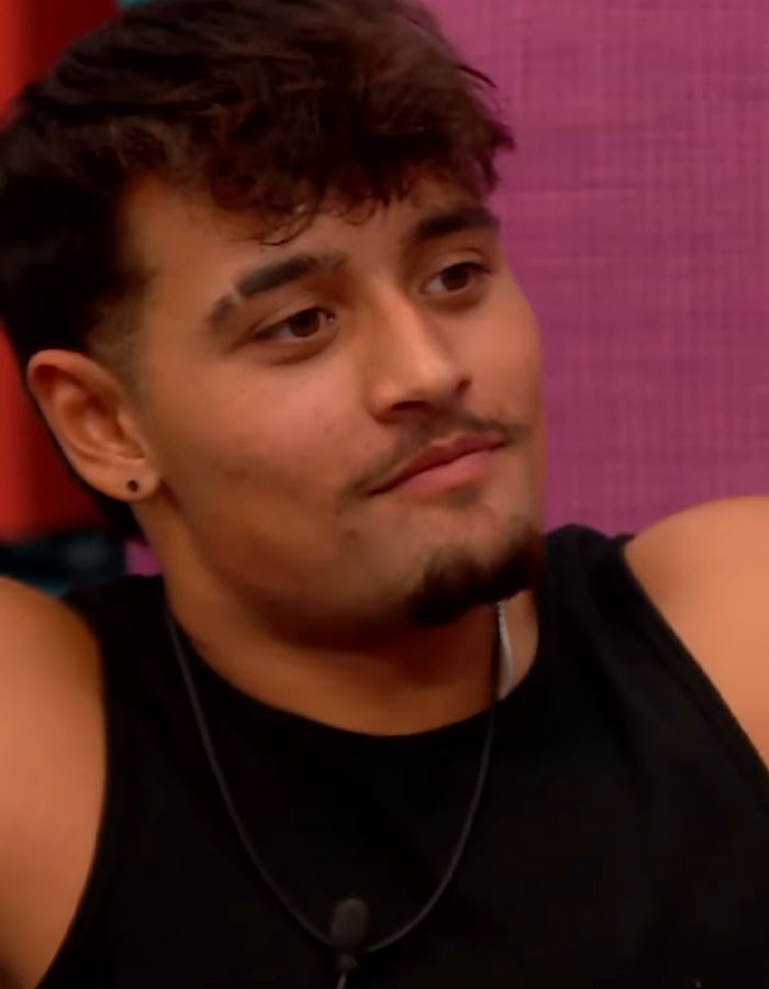Young Big Brother housemate wearing black tank top and necklace looking thoughtful during a tense moment in the house.