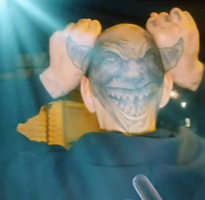 Person showing off an awful tattoo of a demon face on the back of their head under bright light outdoors at night.
