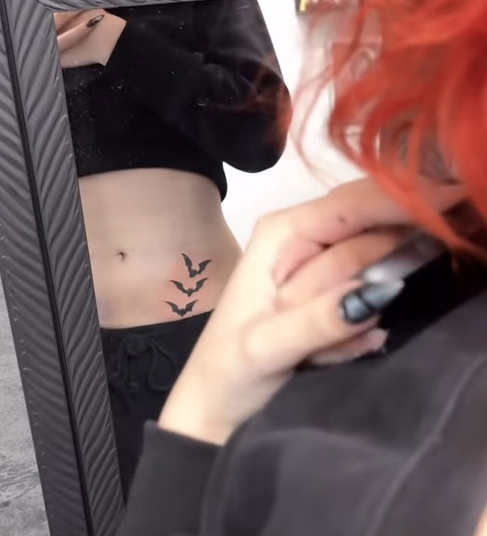 Person with red hair and black nails showing a pelvic tattoo of three bats in a mirror, related to pelvic tattoo controversy. Person with red hair and black nails showing a pelvic tattoo of three bats in a mirror, related to pelvic tattoo controversy.