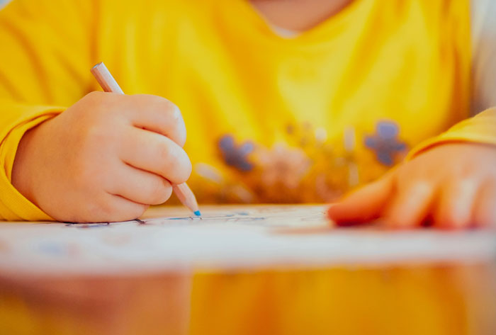 Child in yellow shirt drawing with a colored pencil, illustrating daycare workers dealing with unexpected family secrets.