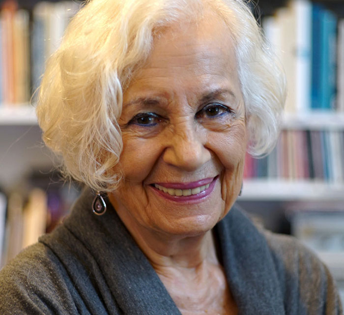 Holocaust survivor Ruth Posner smiling warmly indoors with bookshelves in the background in a close-up portrait.