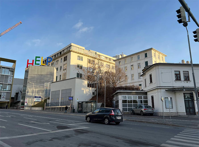 Hospital building with colorful HELP sign on rooftop, cars on street, and clear blue sky in the background.