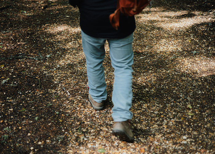 Man walking on a forest path wearing light blue pants and brown boots, surrounded by fallen leaves and dappled sunlight.