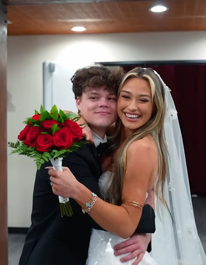 Adult star and YouTube prankster husband in wedding attire, smiling and hugging with bouquet of red roses.