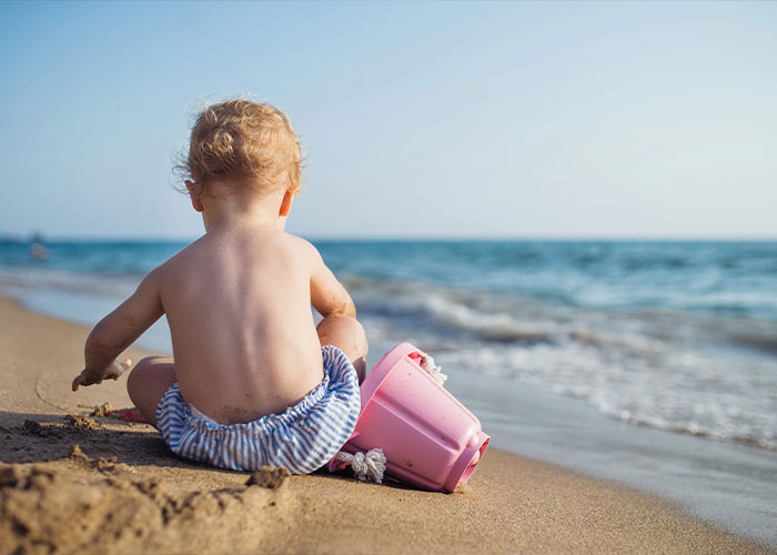6-month-old baby sitting alone on a sandy beach near the water with a pink bucket, major twist case concept.