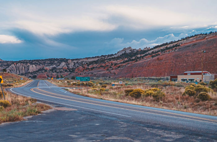 Desert highway curving past rocky cliffs under a cloudy sky, a location from places people wouldn’t revisit list.