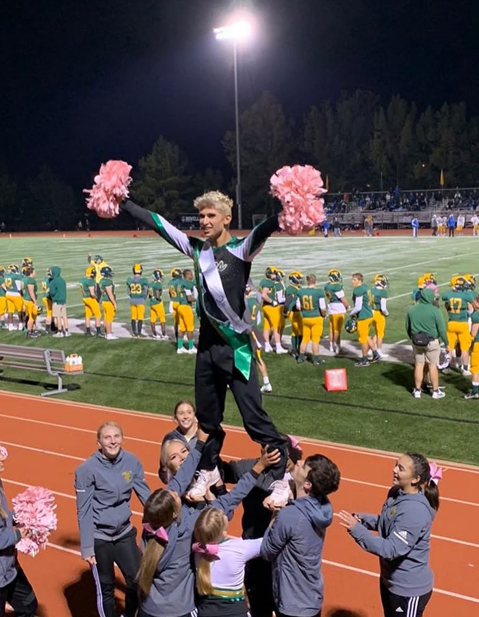 Missouri&rsquo;s first male homecoming queen wearing a sash and cheerleading, lifted by a group on a football field at night.