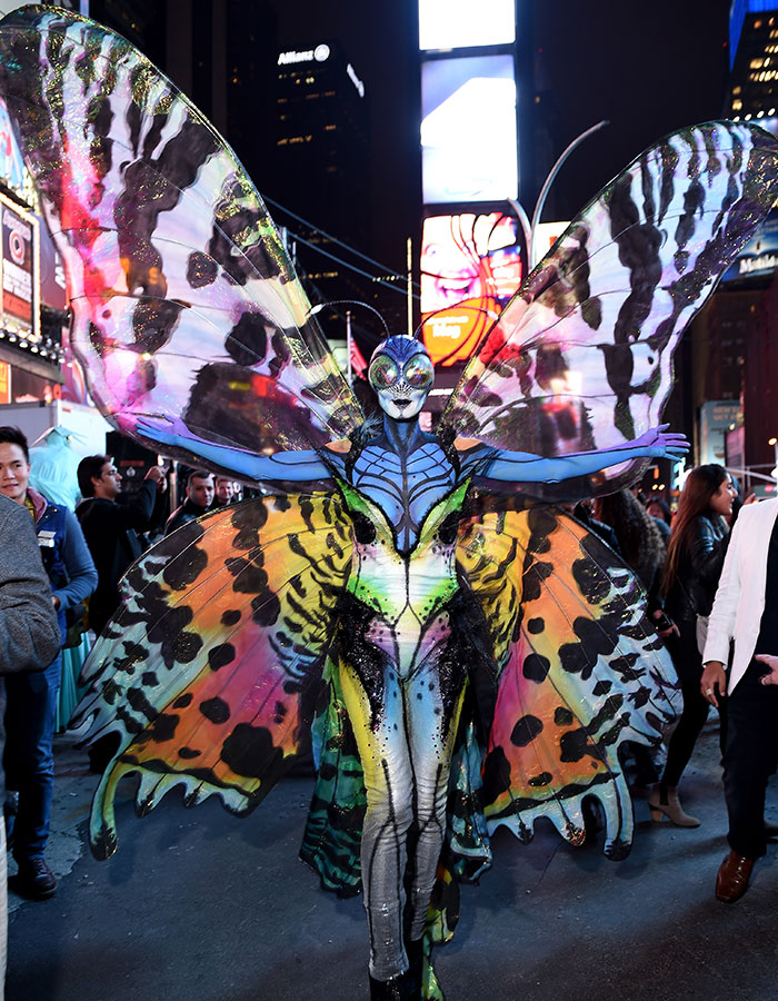 Person in an elaborate butterfly costume with large colorful wings posing on a busy city street at night, Halloween event.