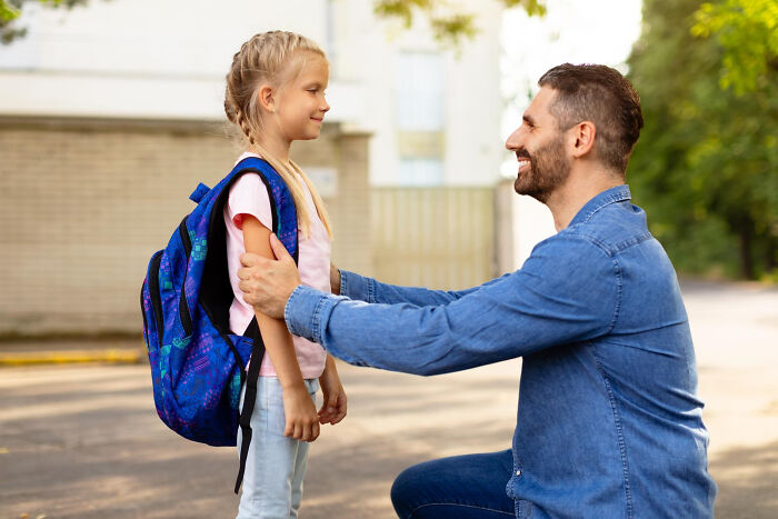 Man kneeling and talking to young girl with backpack outdoors, illustrating unfair relationships due to gender roles.