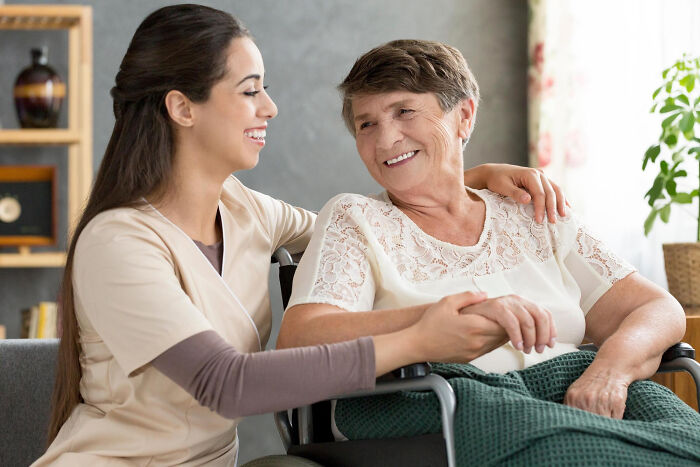 Caregiver smiling and holding hands with a happy elderly woman with dementia in a wheelchair at home.