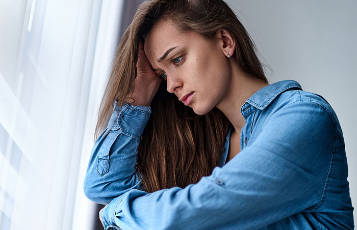 Young woman in a denim shirt looking sad by a window reflecting on bride's father refusing wedding outside American soil