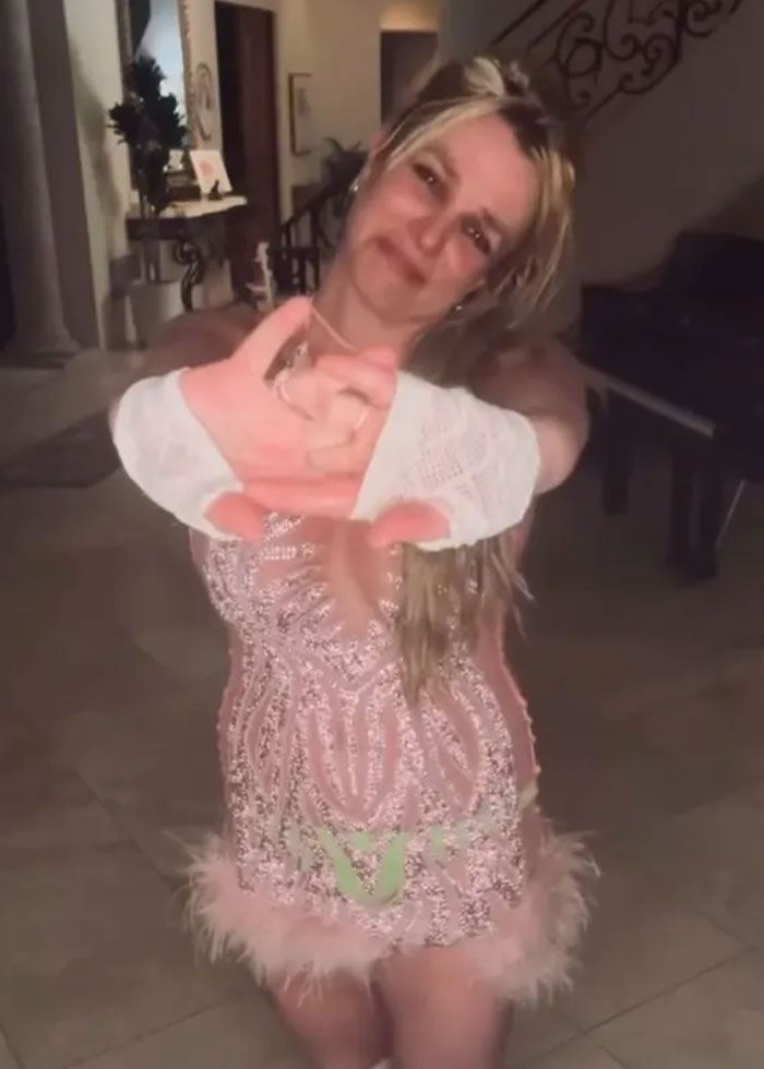 Britney Spears wearing a pink dress with bruises visible on her face while gesturing with her hands indoors.