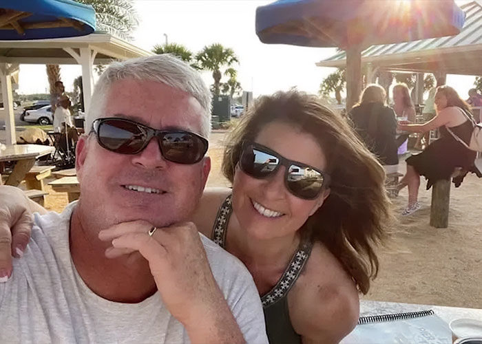 Man and woman wearing sunglasses smiling outdoors at a sunny beachside caf&eacute; with tables and umbrellas.