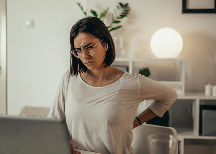 Woman in glasses sitting at a desk holding her back in pain while looking at a laptop, highlighting hidden dangers in household items. Woman in glasses sitting at a desk holding her back in pain while looking at a laptop, highlighting hidden dangers in household items.