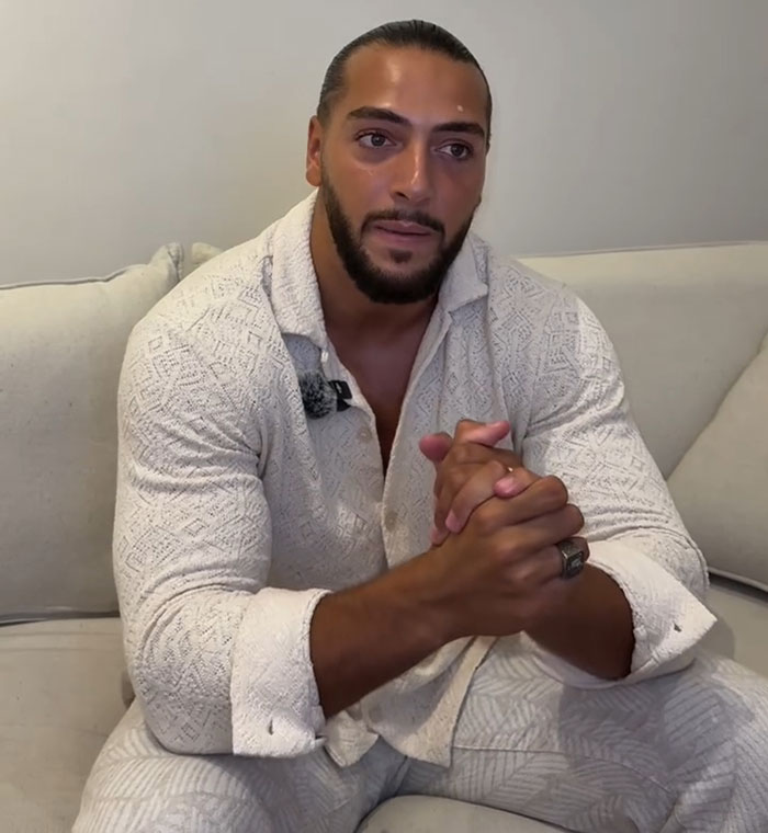 Man in a white textured shirt sitting on a couch, discussing influencer fake syringe attack pranks and their arrest.