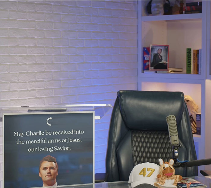 Home office setup with a microphone and a sign referencing Charlie Kirk, related to a controversial homework assignment.