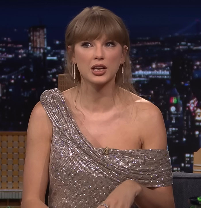 Taylor Swift wearing a sparkling one-shoulder dress, speaking during a nighttime talk show interview.