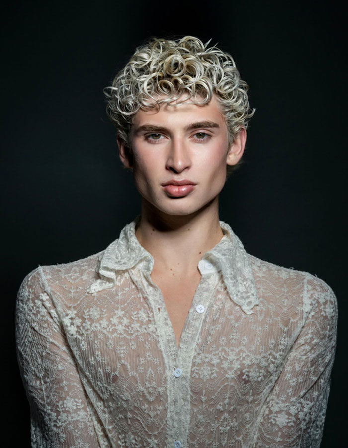 Young man with curly blond hair wearing a sheer lace shirt, representing Missouri&rsquo;s first male homecoming queen in a portrait.