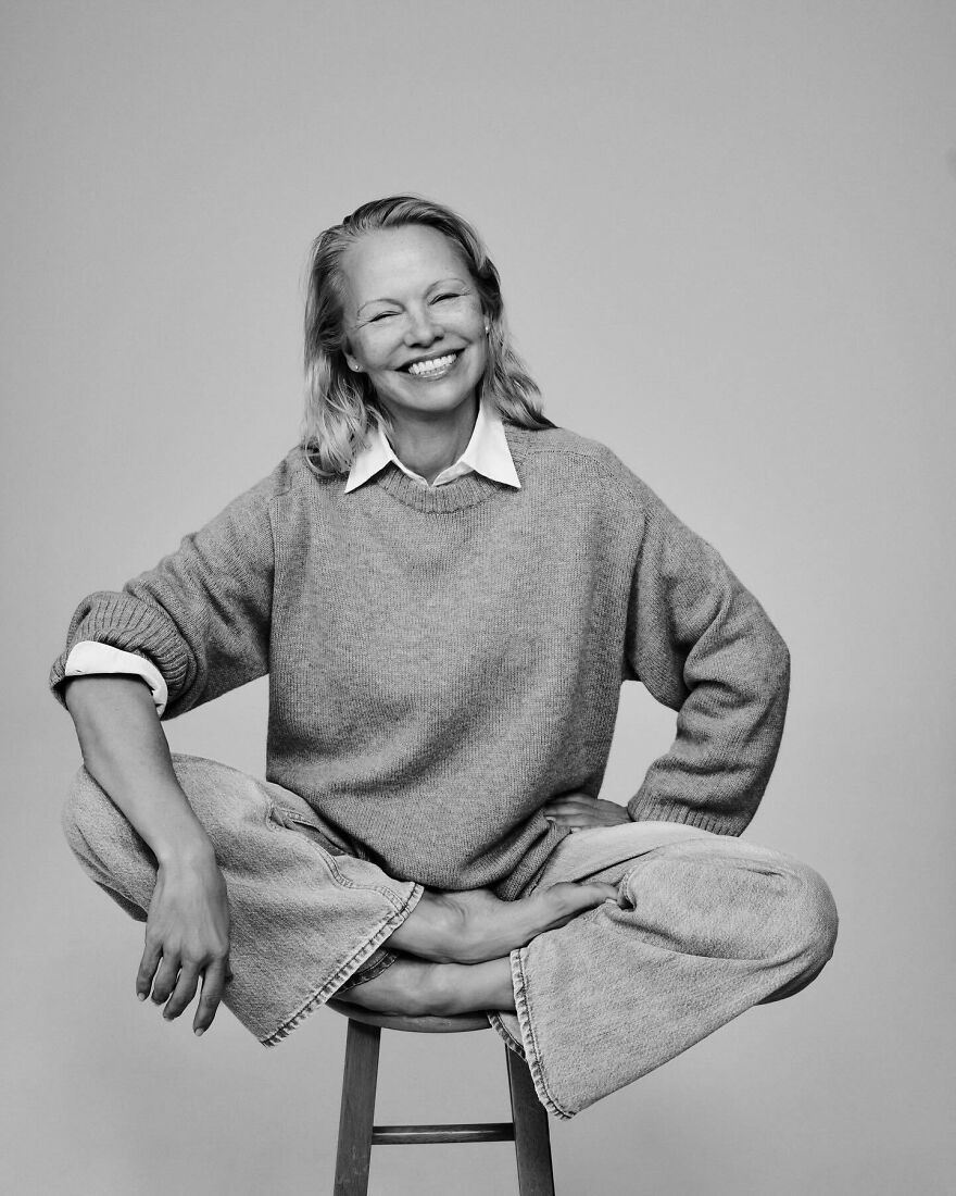 Pamela Anderson smiling seated cross-legged on a stool wearing a sweater and jeans showing confidence and reinvention.