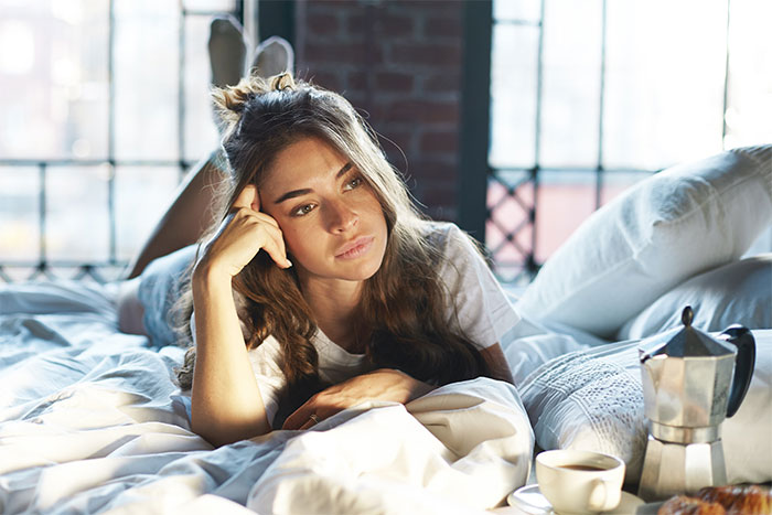 Young woman lying on bed looking thoughtful with coffee and teapot nearby in a cozy bedroom setting.