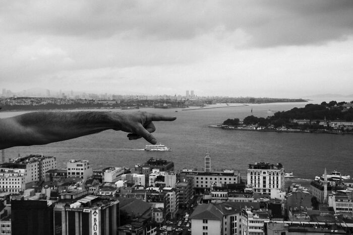 Black and white street photography shot of a hand pointing over a cityscape by a river on a cloudy day.